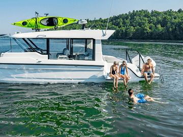 A Cruisers Yachts 38 VTR on calm water with four people relaxing on and around it, including two sitting on the side platform and one swimming in the foreground. A bright green kayak is mounted on top of the boat, and a tree-lined shore is visible in the background.