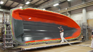 A large boat hull section being sprayed inside a factory workshop, with a worker standing beside it.